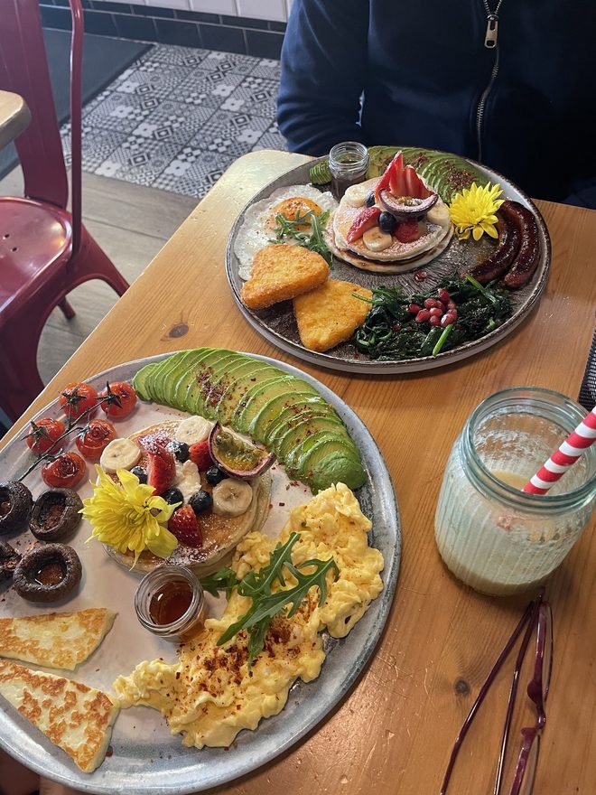 Two platters of scrambled eggs, avocado, halloumi and pancakes with a milkshake, seen across the table